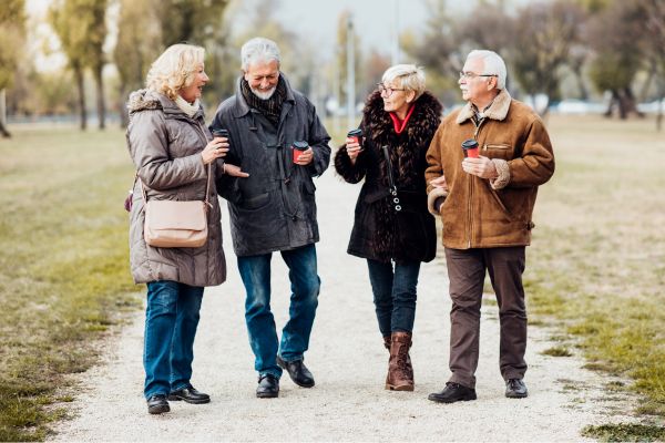 Vier Senioren gehen gemeinsam spazieren im Park, unterhalten sich und halten Becher in der Hand.